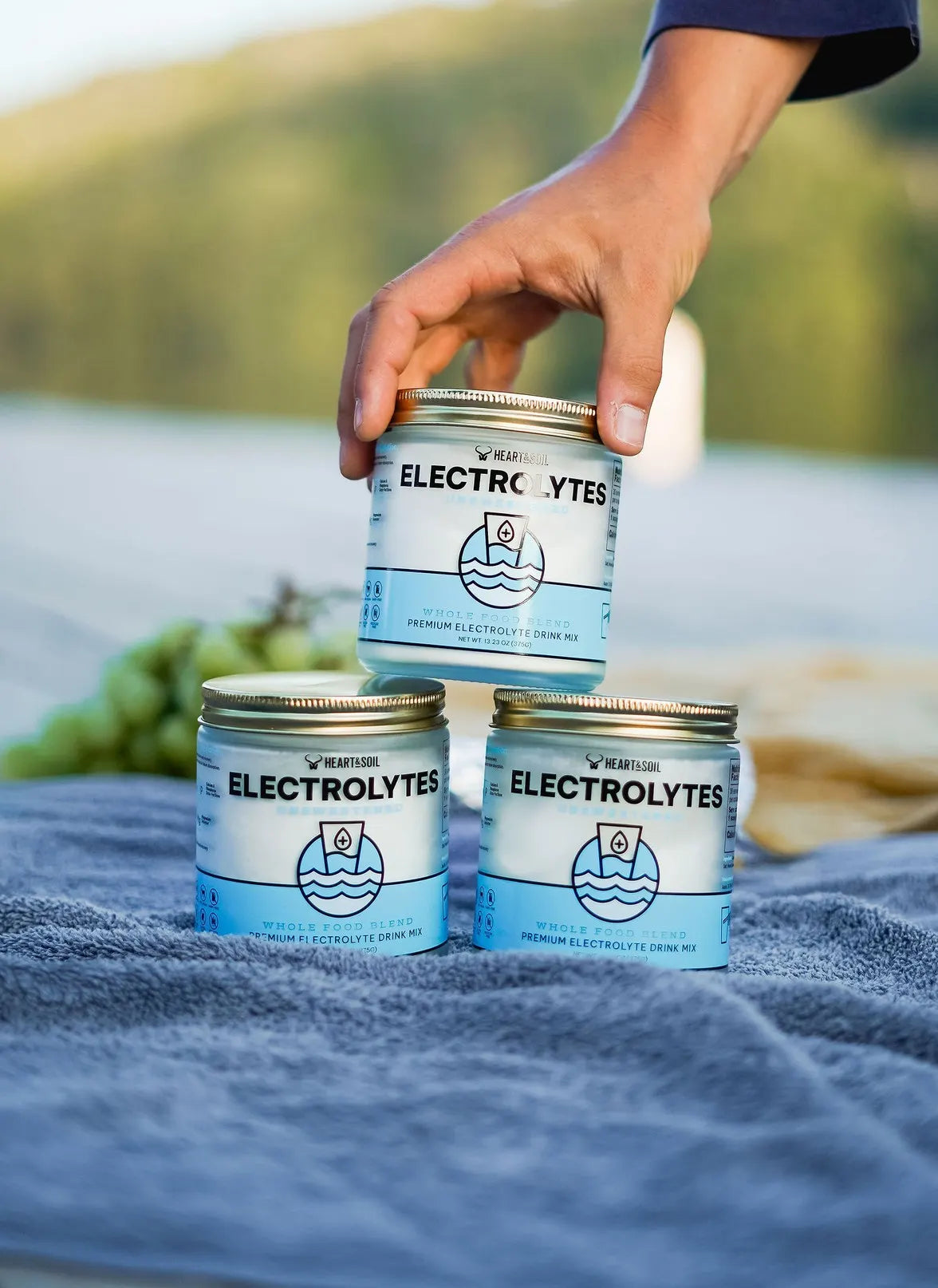 A hand stacking Heart & Soil Electrolytes jars on a blanket outdoors
